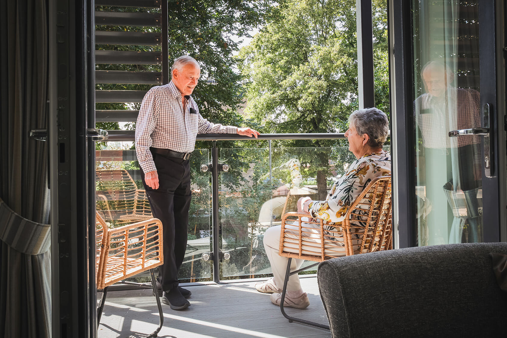 Maura and David on their balcony, Maura is sat on an arm chair and David is stood resting his arm on the handrail.