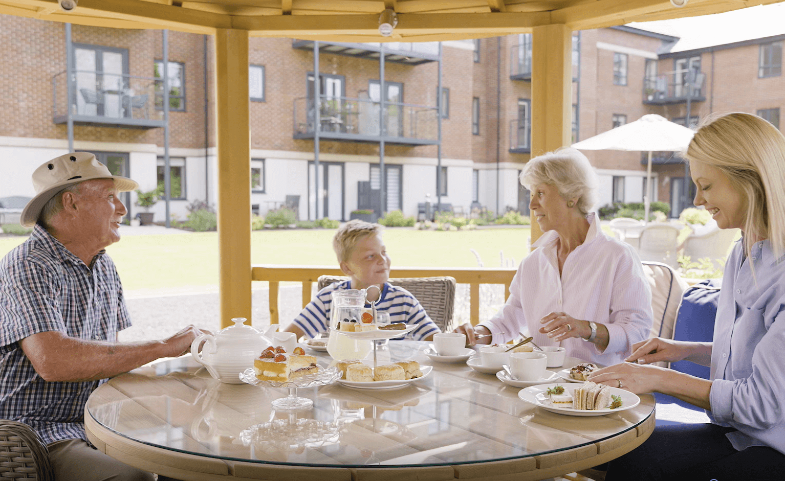 Retired couple enjoying afternoon tea in a summer house within landscaped gardens with their daughter and grandson.