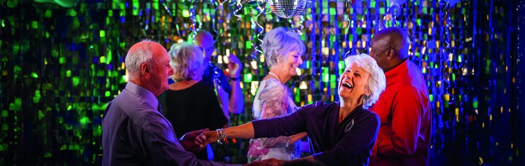 A group of older adults are happily dancing under a disco ball, surrounded by green and blue shimmering decorations, smiling and enjoying the lively atmosphere..jpg