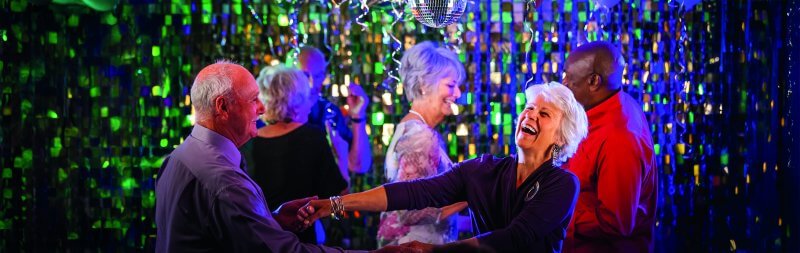 A group of older adults are happily dancing under a disco ball, surrounded by green and blue shimmering decorations, smiling and enjoying the lively atmosphere..jpg